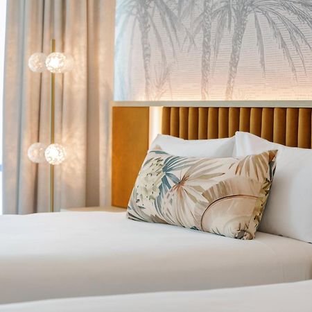 Amora Hotel בריסביין מראה חיצוני תמונה The photo shows a stylish hotel room featuring two neatly made beds with white linens. Each bed has several pillows, one of which has a decorative cover with a tropical plant pattern. Behind the beds is a modern headboard, and the wall is decorated w