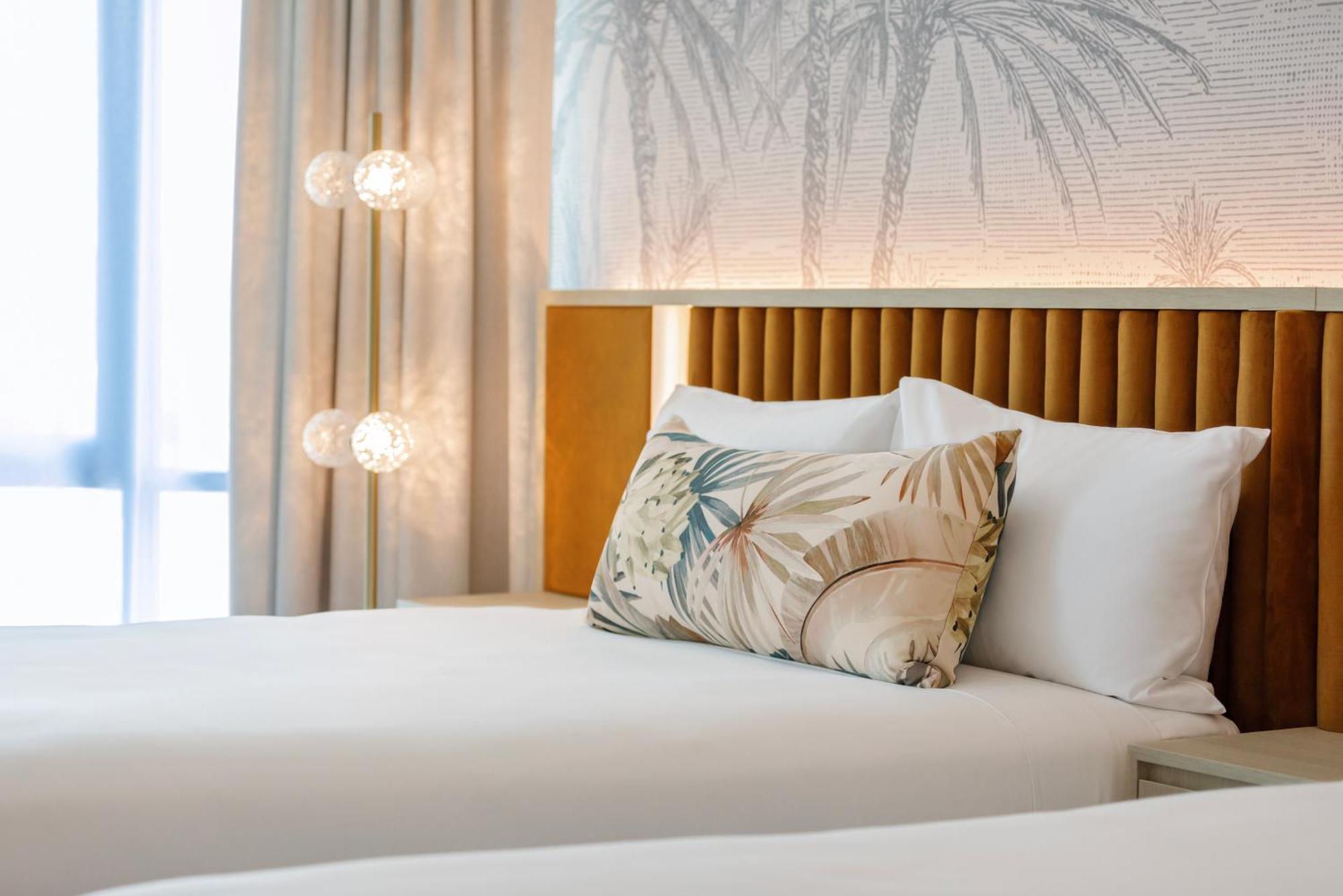 Amora Hotel בריסביין מראה חיצוני תמונה The photo shows a stylish hotel room featuring two neatly made beds with white linens. Each bed has several pillows, one of which has a decorative cover with a tropical plant pattern. Behind the beds is a modern headboard, and the wall is decorated w