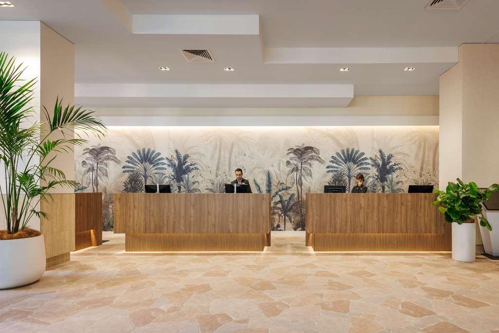 Amora Hotel בריסביין מראה פנימי תמונה The photo shows a modern reception area with two wooden reception desks. Behind the desks, there is a decorative wall featuring a large mural of tropical plants and palm trees, creating a serene atmosphere. The space is well-lit with overhead lights,